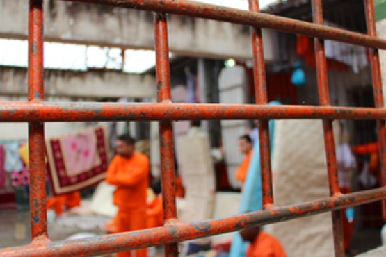 Fotografia de penitenciária com o foco nas grades e ao fundo, em desfoque, estão os detentos perto de um varal com cobertores pendurados