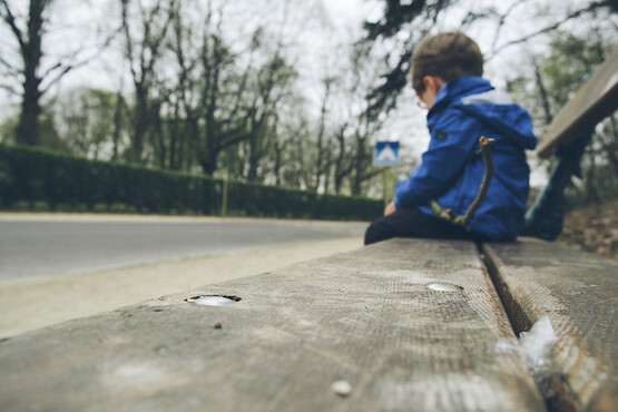 Foto ilustrativa de uma criança de casaco azul sentada sozinha em um banco de madeira. A imagem foi capturada em um ângulo de baixo para cima, pegando o banco em primeiro plano e a criança ao fundo.