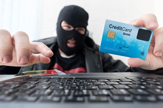 Foto ilustrativa de homem branco de balaclava segurando e olhando os dados de um cartão de crédito enquanto digita em um notebook.