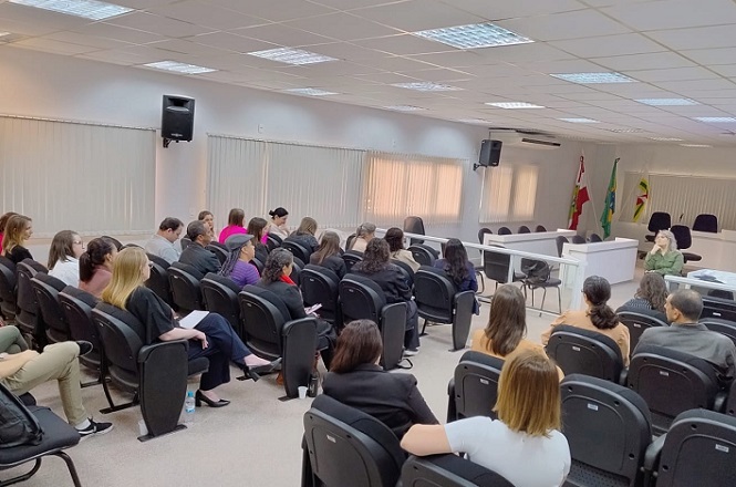 Grupo de alunos em sala de aula escuta formadora, que veste blusa verde.