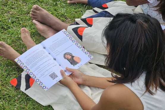 Jovem índia, sentada na grama da aldeia, com um exemplar piloto da futura cartilha da Lei Maria da Penha já traduzida para a língua Xokleng