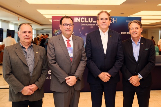 A imagem mostra quatro homens posando para uma foto em um ambiente interno. Todos estão usando ternos. O fundo parece ser um local de evento, a iluminação é clara e o ambiente parece ser um salão de conferências ou algo similar.