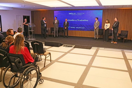 A imagem mostra um evento em um auditório. No lado esquerdo, há uma pessoa em uma cadeira de rodas e outras pessoas sentadas próximas a ela. No centro, há um palco elevado com quatro pessoas em pé, duas delas usando ternos. No fundo do palco, há uma tela grande com texto em azul, mas o conteúdo do texto não está legível na imagem. No lado direito do palco, há um púlpito com uma pessoa falando ao microfone. O ambiente parece ser formal, possivelmente uma conferência ou cerimônia.