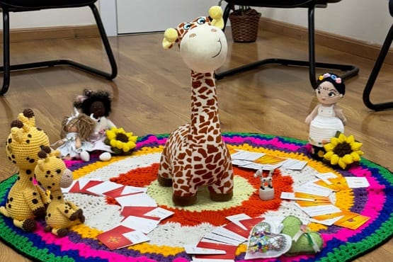 Girafa de pelúcia no centro de um círculo feito de tricô. Em volta da pelúcia também existem outros brinquedos e cartas.  
