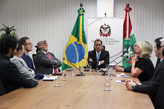 Autoridades do TJSC sentadas em volta de uma mesa. Ao fundo estão duas bandeiras e um banner do Tribunal de Justiça do Estado de Santa Catarina.