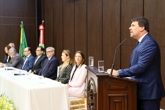 No lado direito da imagem está um homem, que está atrás do púlpito, falando em um microfone e, na esquerda, está a mesa de autoridades compostas por três mulheres e cinco homens. Ao fundo estão duas bandeiras.