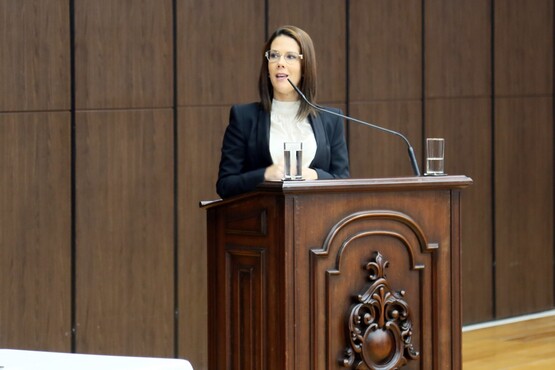 Em um púlpito, uma mulher de óculos, que veste uma blusa branca e uma blazer preto, fala no microfone.