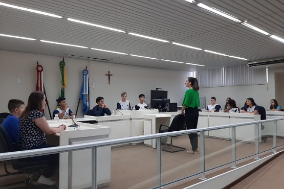Vereadores mirins sentados em mesas que juntas formam um semicírculo na sala das sessões do Tribunal do Júri na comarca de Videira. Ao centro da está uma mulher de blusa verde olhando para as crianças e ao fundo estão três bandeiras.