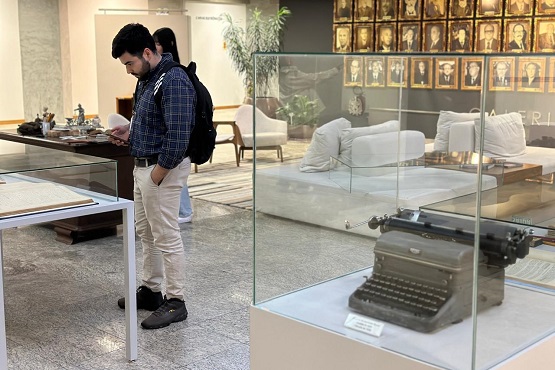 Em primeiro plano está uma máquina de escrever dentro de uma redoma de vidro quadrada e, ao fundo, um homem olha outra parte da exposição.