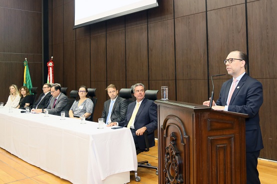 No lado direito da imagem, um homem engravatado de óculos fala no microfone em um púlpito. À esquerda, estão sete pessoas sentadas na mesa de autoridades. 