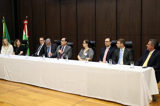 Mesa de autoridades composta por três mulheres e seis homens vestidos de modo formal. Ao fundo existem duas bandeiras.