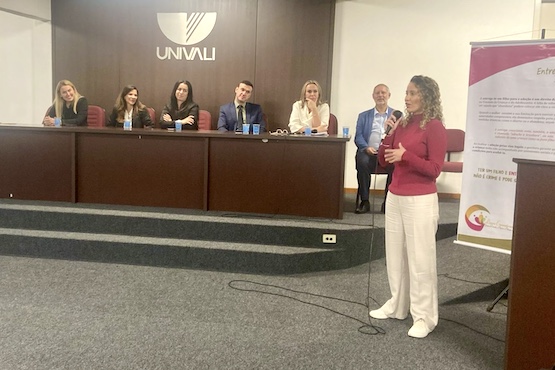 Pessoas de costas para a imagem acompanham a fala de um homem engravatado, que está ao centro da foto. Ao fundo, está uma mesa de autoridades.