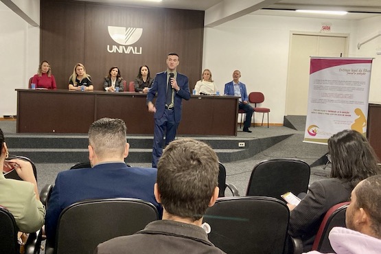 Pessoas de costas para a imagem acompanham a fala de um homem engravatado, que está ao centro da foto. Ao fundo, está uma mesa de autoridades.