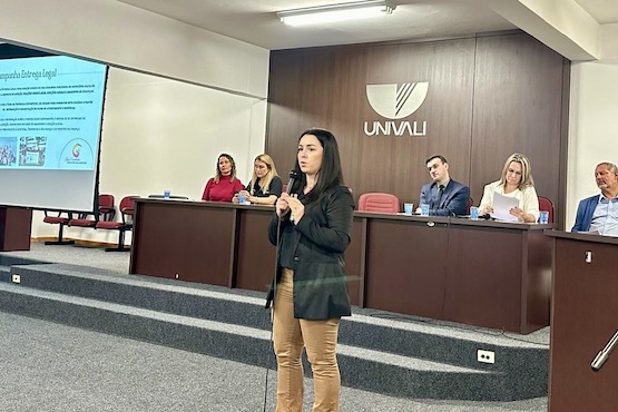 Foto de uma mulher, que está localizada ao centro da imagem, falando no microfone. Ao fundo está uma mesa de autoridades.