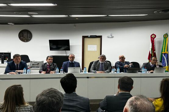 A imagem mostra uma reunião formal com seis homens sentados atrás de uma mesa comprida. Eles estão vestidos com ternos e gravatas. Na mesa, há garrafas de água, copos e alguns laptops. Atrás deles, na parede, há uma televisão, uma porta e bandeiras do Brasil e de Santa Catarina. Na frente da mesa, há várias pessoas sentadas, de costas para a câmera, assistindo à reunião.