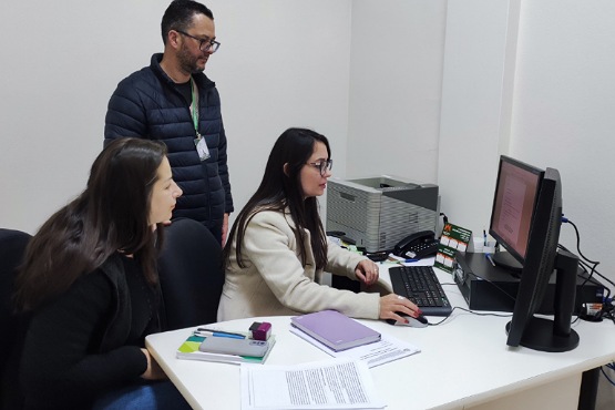 A imagem mostra três pessoas em um escritório. Duas mulheres estão sentadas em frente a um computador, enquanto um homem está em pé atrás delas. A mulher à direita está digitando no teclado e olhando para a tela do computador. Ela usa óculos e um casaco claro. A mulher à esquerda está olhando para a tela do computador e tem cabelos longos e escuros. O homem em pé está usando óculos, uma jaqueta preta e um crachá pendurado no pescoço. Na mesa, há vários papéis, um caderno roxo, um estojo de canetas e um telefone fixo. Ao fundo, há uma impressora e mais papéis empilhados.