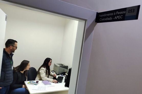 A imagem mostra uma sala de atendimento com três pessoas. Duas mulheres estão sentadas em uma mesa branca, uma delas digitando em um computador e a outra observando. Um homem está em pé ao lado delas, também observando. Na parede ao lado da porta, há uma placa preta com a inscrição "Atendimento à Pessoa Custodiada - APEC" e o número "053". A sala é simples, com paredes brancas e um ambiente de escritório.