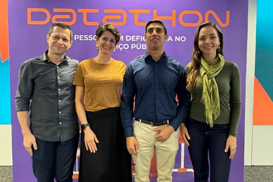 A imagem mostra quatro pessoas posando para uma foto em frente a um banner roxo com a palavra "Datathon" escrita em laranja. Abaixo da palavra "Datathon", está escrito "Pessoas com Deficiência no Serviço Público". Da esquerda para a direita, há um homem de óculos com uma camisa azul escura, uma mulher de cabelo curto e castanho usando uma blusa laranja e uma saia preta, um homem de cabelo curto e escuro usando uma camisa azul escura e calça clara, e uma mulher de cabelo longo e castanho usando uma blusa verde e um cachecol verde claro. Todos estão sorrindo.