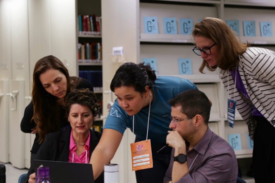 A imagem mostra cinco pessoas em um ambiente de escritório ou biblioteca, concentradas em torno de um laptop. Quatro mulheres e um homem estão presentes. Uma mulher de camiseta azul está apontando para a tela do laptop, enquanto as outras pessoas observam atentamente. Ao fundo, há estantes com livros e pastas organizadas. Todos parecem estar envolvidos em uma discussão ou apresentação.