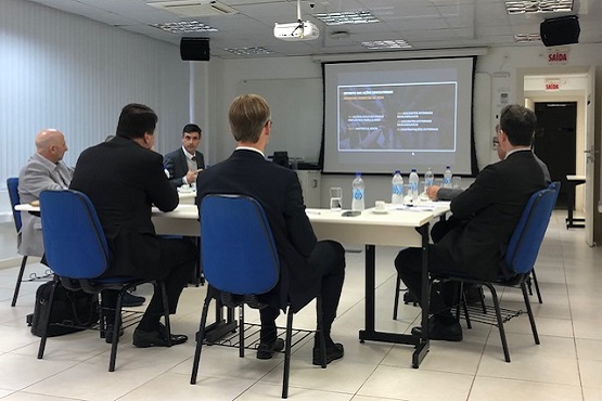 A imagem mostra uma sala de reuniões com cinco pessoas sentadas em cadeiras azuis ao redor de uma mesa. Quatro delas estão de costas para a câmera e uma está de frente, parecendo estar apresentando algo. Na parede à frente, há uma tela de projeção com uma apresentação exibida. A sala tem um ambiente corporativo, com piso claro e teto com iluminação embutida.