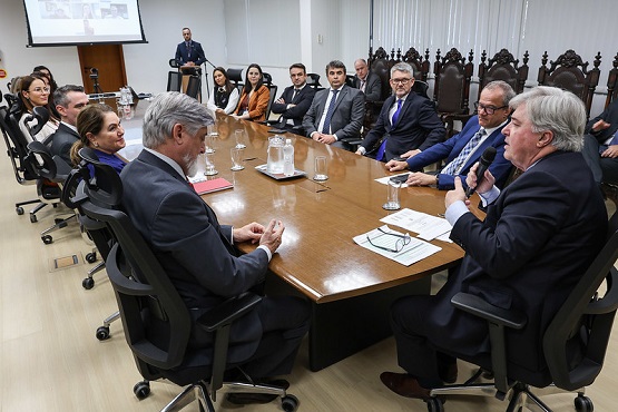 A imagem mostra uma reunião formal em uma sala de conferências. Há uma mesa grande de madeira no centro, em formato oval, ao redor da qual estão sentadas várias pessoas, a maioria delas usando trajes formais como ternos e gravatas. Na parte mais próxima da câmera, à direita, um homem de cabelos grisalhos está falando e gesticulando com a mão direita. Ele parece estar liderando a reunião ou fazendo uma apresentação. Ao seu lado esquerdo, está outro homem de cabelos. Ao fundo, há mais pessoas sentadas, algumas mulheres e homens, todos prestando atenção na reunião. Na parede ao fundo, há uma tela de projeção com uma imagem exibida, e um homem de terno está em pé ao lado da tela, possivelmente um assistente. A sala tem um ambiente formal, com cadeiras de escritório ao redor da mesa e cadeiras de madeira esculpidas ao fundo, sugerindo um ambiente de alta importância ou oficial.