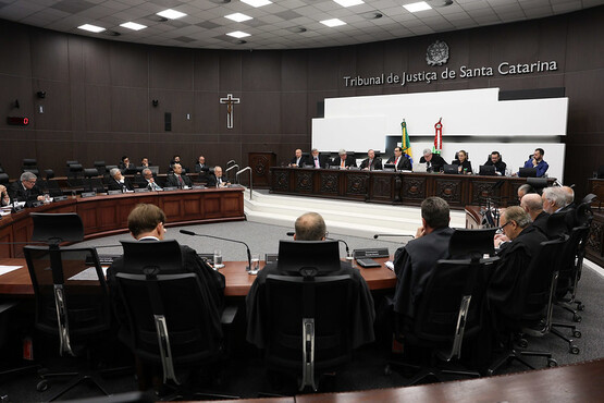 A imagem mostra uma sala de tribunal no Tribunal de Justiça de Santa Catarina. A sala é ampla e possui um grande número de cadeiras dispostas em semicírculo. Na parte frontal, há uma bancada elevada onde estão sentados vários juízes ou autoridades judiciais. Atrás deles, na parede, está escrito "Tribunal de Justiça de Santa Catarina" e há um brasão acima do texto. Também é possível ver um crucifixo na parede à esquerda. As bandeiras do Brasil e de Santa Catarina estão posicionadas atrás da bancada dos juízes. A sala está bem iluminada e há várias pessoas sentadas, aparentemente participando de uma sessão ou audiência.