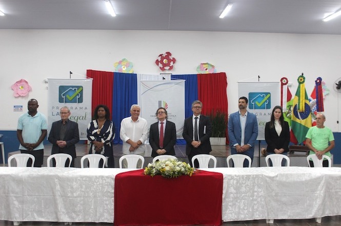 A imagem mostra um evento formal em um salão decorado. Há oito pessoas de pé, entre elas o Des. Selso de Oliveira e demais autoridades da solenidade, todas atrás de uma mesa comprida coberta com toalhas brancas e um arranjo de flores. No fundo, há cortinas coloridas e banners com o texto "Programa Lar Legal". Há também bandeiras. As pessoas estão vestidas formalmente.