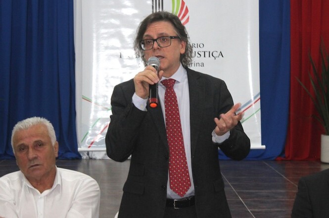 A imagem mostra o Des. Selso de Oliveira falando ao microfone em um evento formal. Ele veste um terno preto, camisa azul clara e uma gravata vermelha. Atrás dele, há uma cortina e um banner branco com o logotipo do PJSC, onde se lê "Ministério Público de Justiça de Santa Catarina". Ao lado esquerdo dele, um homem de camisa branca está sentado.