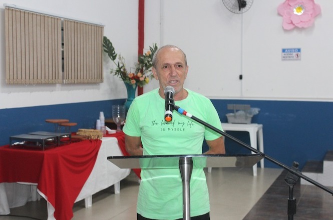 Um homem com camiseta verde está falando ao microfone, com as mãos para trás e de pé atrás de um púlpito de metal e vidro. O local é simples, com uma mesa ao fundo coberta com toalha, além de arranjos decorativos. A parede é branca com detalhes em azul.