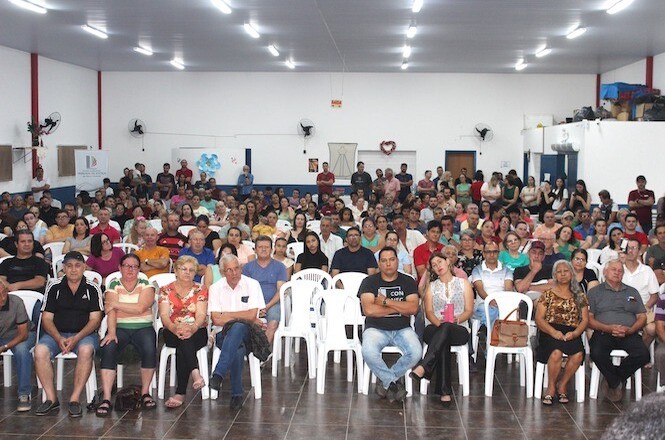 A imagem mostra uma fotografia de um grupo numeroso de pessoas sentadas em cadeiras brancas em um salão amplo, assistindo a um evento. As cores predominantes do local são tons de cinza, branco e vermelho.