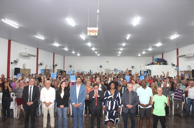 A imagem mostra uma fotografia de um grupo numeroso de pessoas em pé em frente à suas cadeiras em um salão amplo, pousando para a fotografia, alguns estão levantando e mostrando papéis ou documentos. As cores predominantes do local são tons de cinza, branco e vermelho.