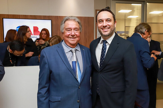 Foto colorida: duas pessoas de terno posam juntas, sala institucional interna, evento formal, registrando encontro em azul, branco e bege 