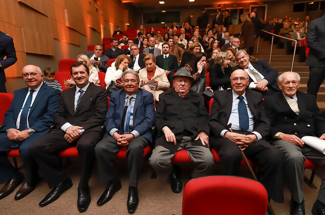 Foto colorida: autoridades sentadas em auditório com poltronas vermelhas, ambiente institucional interno, evento formal, acompanhando solenidade em vermelho, preto e dourado