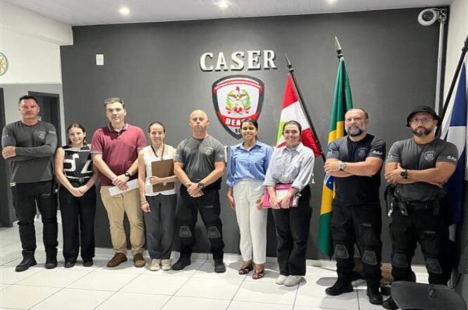 Foto colorida: grupo alinhado em sala do CASER, ambiente institucional, visita oficial, registrando encontro em cinza, verde e branco