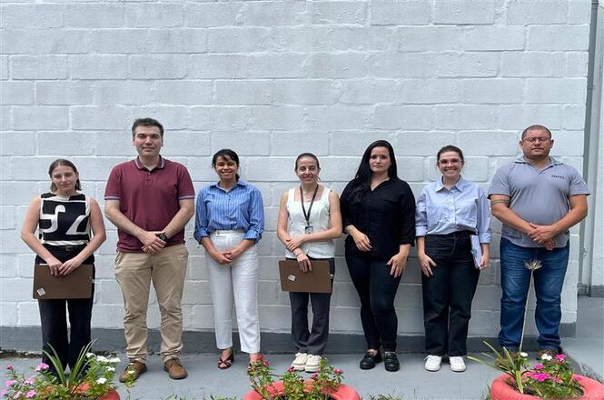 Foto colorida: grupo alinhado em área externa institucional, muro claro ao fundo, visita técnica, posando para registro 