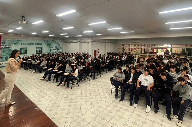 Foto colorida: palestrante à frente de grande público, auditório escolar amplo, ação educativa, conduzindo palestra em branco, cinza e verde