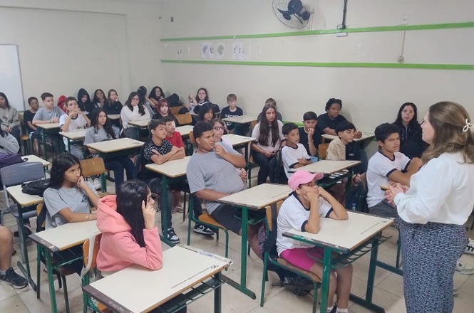 Foto colorida: estudantes sentados em carteiras, sala de aula interna, ação educativa, acompanhando orientação em branco, verde e bege