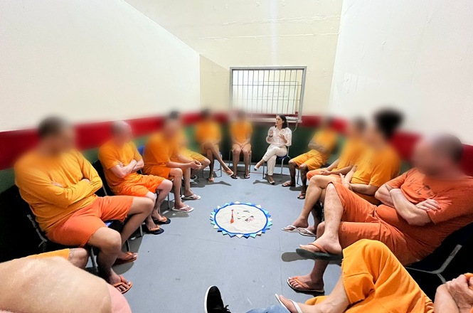 Foto colorida: grupo de detentos sentados em círculo em sala interna, atividade restaurativa institucional, dialogando em laranja, branco e cinza