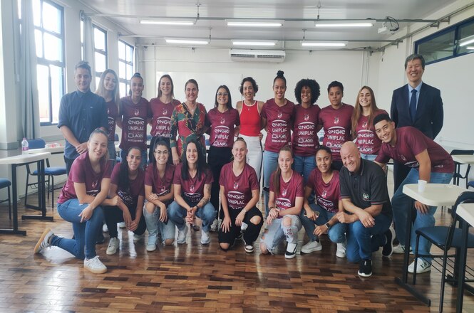 Foto horizontal: grupo de mulheres posa em sala após atividade educativa; jovens com camisetas do time Leoas da Serra; ambiente claro