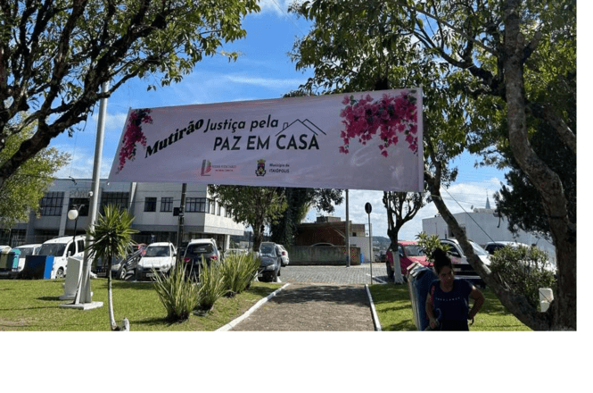 Foto colorida: faixa “Mutirão Justiça pela Paz em Casa” em jardim urbano, área externa do fórum, ação institucional, sinalizando atendimento em branco, rosa e verde