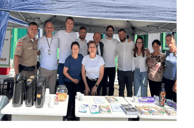 Foto colorida: grupo reunido sob tenda com materiais informativos, área externa urbana, ação comunitária, orientando público em branco, verde e azul