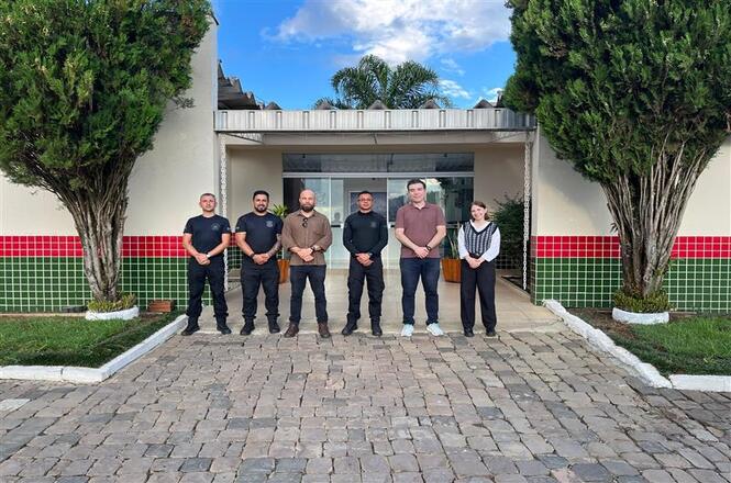Foto colorida: grupo alinhado em frente à Penitenciária Industrial de Curitibanos, área externa institucional, visita oficial, registrando equipe