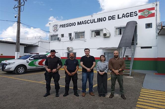 Foto colorida: grupo alinhado em frente ao Presídio Masculino de Lages, área urbana externa, visita institucional, posando para registro