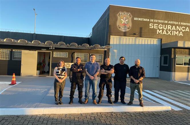 Foto colorida: grupo alinhado em frente à Unidade de Segurança Máxima de São Cristóvão do Sul, área externa institucional, visita oficial, registrando equipe