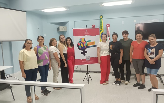 Foto horizontal: grupo de mulheres posa com banner do Programa Indira em sala; ambiente comunitário