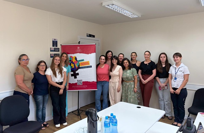 Foto horizontal: grupo de mulheres posa com banner do Programa Indira em sala administrativa; ambiente institucional