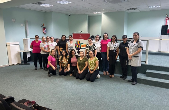 Foto horizontal: grupo de mulheres posa com banner do Programa Indira em auditório; ambiente institucional