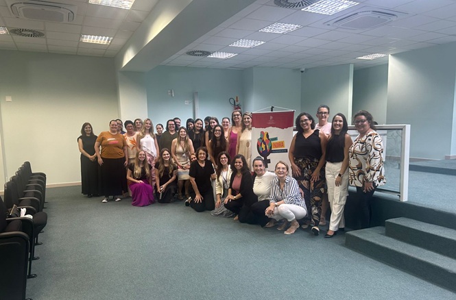Foto horizontal: grupo de mulheres posa com banner do Programa Indira em auditório; ambiente institucional; tons verde, bege e branco.