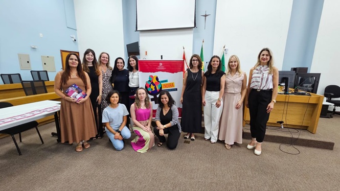 Foto horizontal: grupo de mulheres posa com banner do Programa Indira em sala institucional; ambiente formal; tons bege, preto e vermelho.