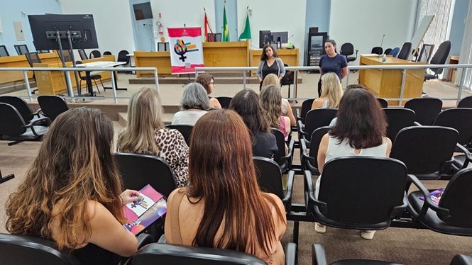 Foto horizontal: mulheres assistem apresentação do Programa Indira em auditório; palestrantes à frente; ambiente institucional; tons bege, verde e branco.